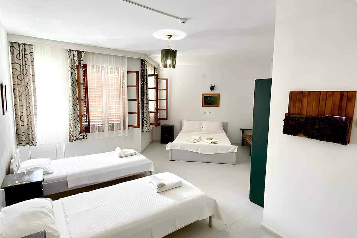 A clean, well-lit hotel room with two single beds, a small desk, a chair, a wardrobe, and a window with curtains.