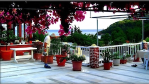 A terrace with potted plants, a white table, and a view of the sea. - Akdeniz Pansiyon (Koycegiz)