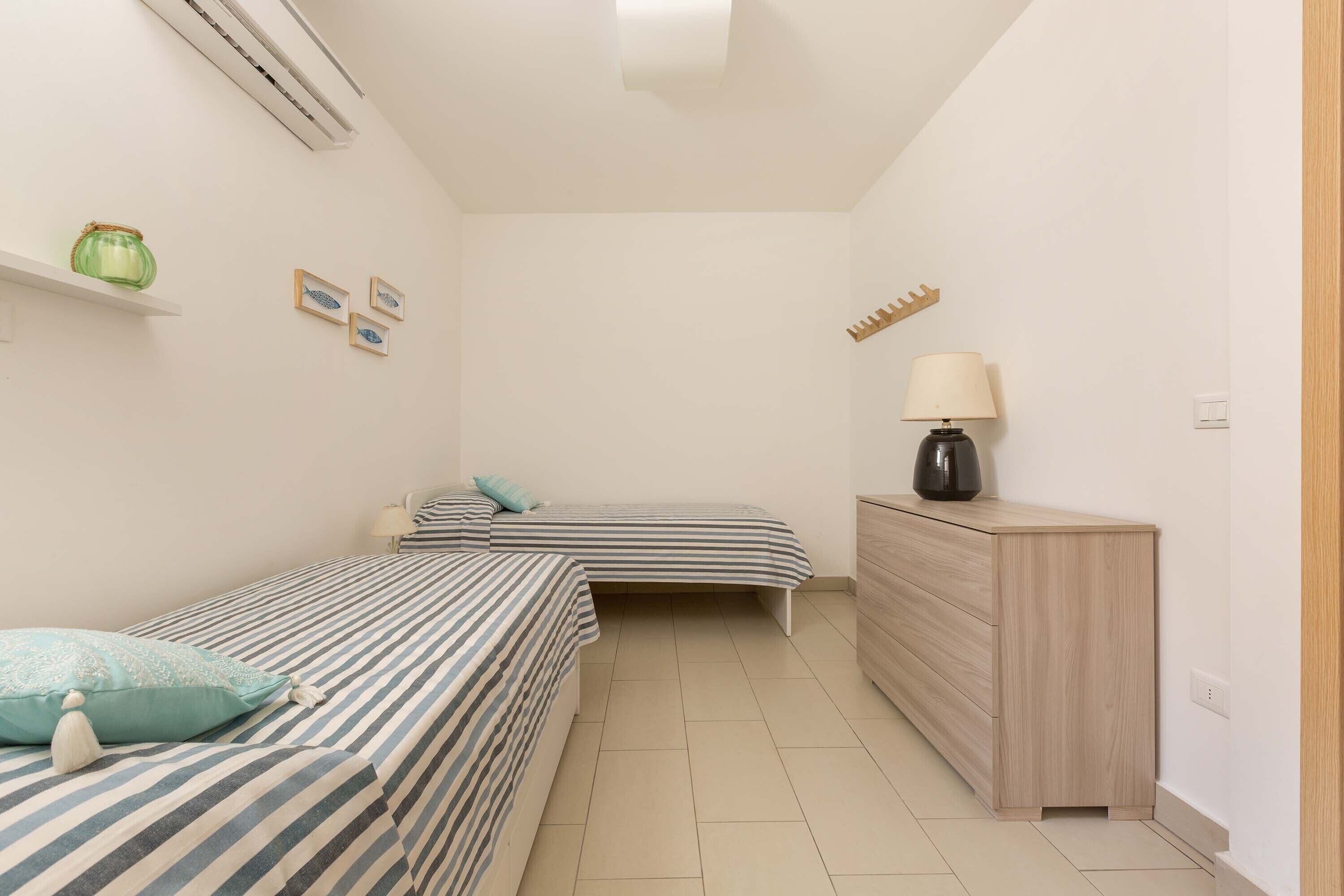 A small, clean room with two single beds, a wooden dresser, a lamp, and a shelf with a jar.