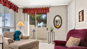 A living room with a beige sofa, a red armchair, a lamp, a mirror, and a sliding glass door leading to a balcony.