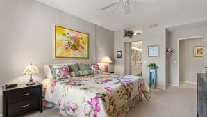 A bedroom with a floral bedspread, a nightstand with a lamp, a ceiling fan, and a large painting on the wall.