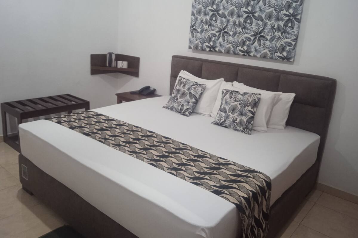 A bedroom with a large bed, a headboard, a bedside table, a nightstand, a wall-mounted shelf, and a wall art piece.