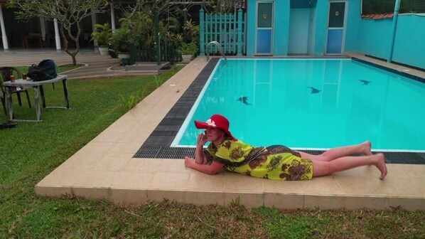 A person lying on a concrete platform by a swimming pool with a tiled deck and a small table with chairs.
