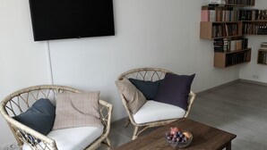 Un salon avec deux fauteuils en osier, une table basse en bois sur laquelle sont disposés des fruits, un téléviseur à écran plat et une bibliothèque remplie de livres.