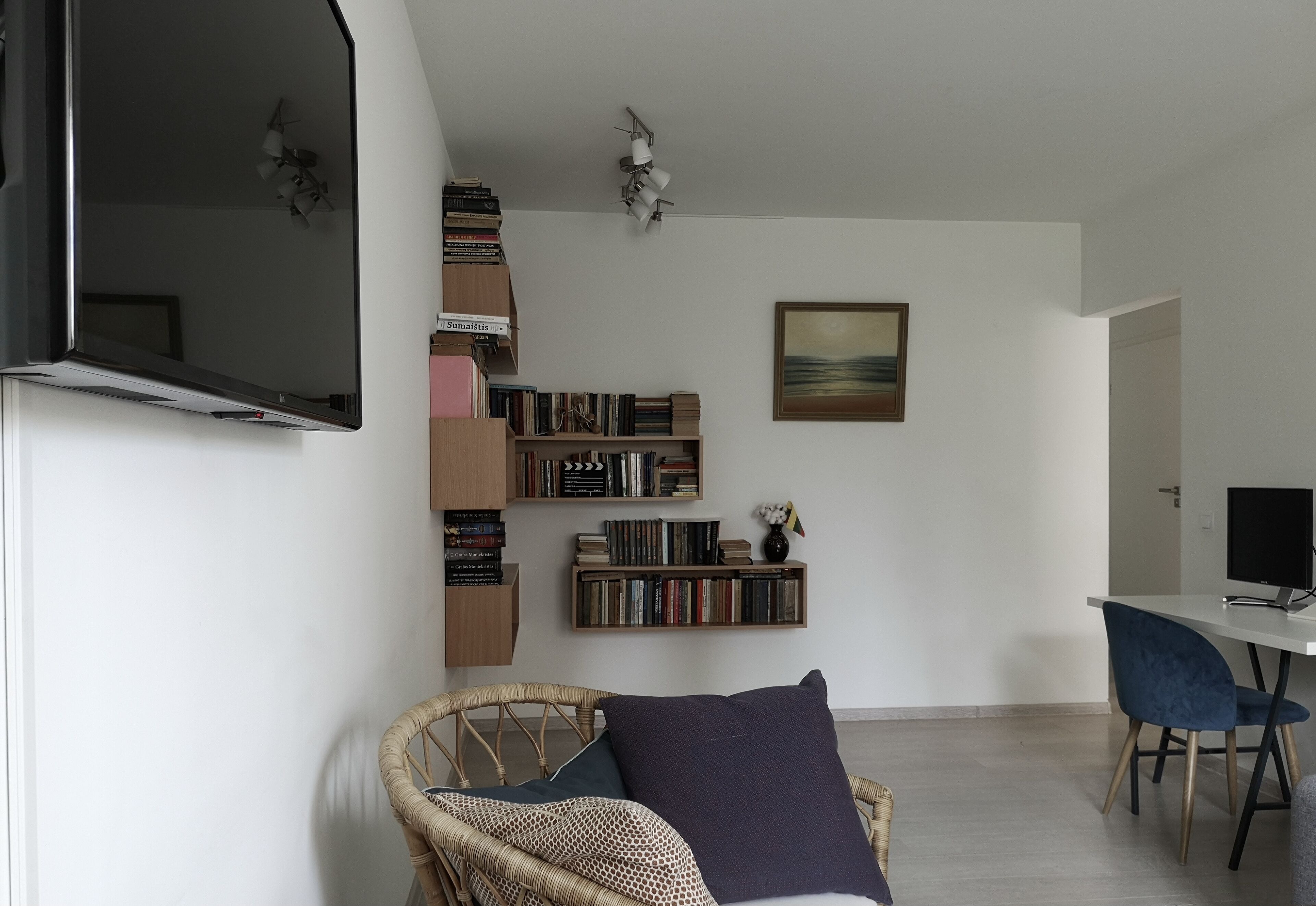 A modern living room with a flat-screen TV, a bookshelf, a painting, and a dining area with a chair and table.