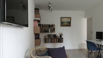 A modern living room with a flat-screen TV, a bookshelf, a painting, and a dining area with a chair and table.