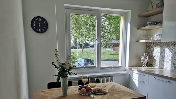 Une cuisine équipée d’une table à manger, de chaises, d’une horloge, d’une fenêtre donnant sur l’extérieur et d’étagères.