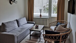Un salon avec un canapé gris, une table basse en bois, un fauteuil en osier et une grande fenêtre avec des rideaux.