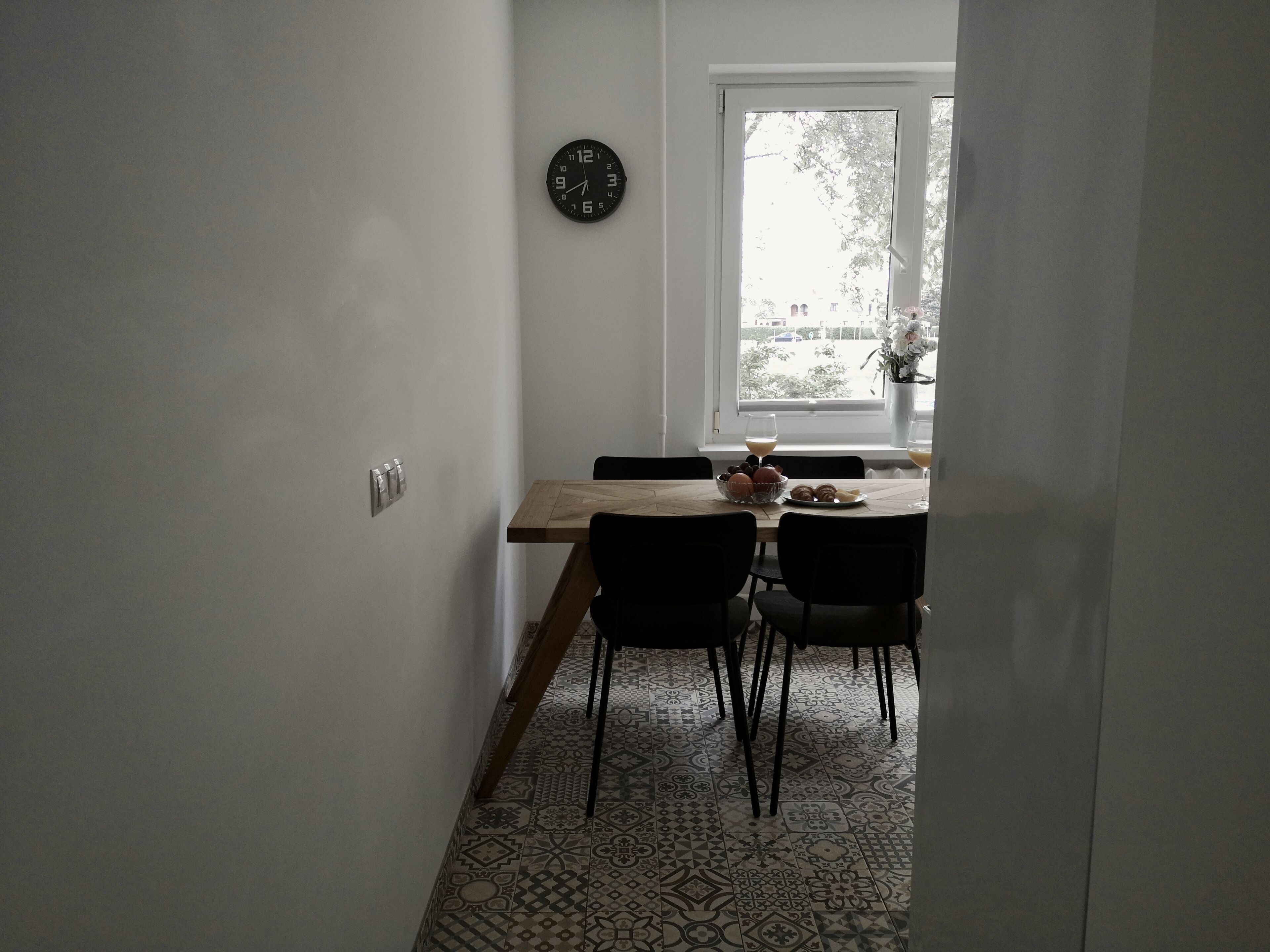 Un couloir étroit mène à une salle à manger où se trouvent une table en bois, des chaises et une horloge au mur.