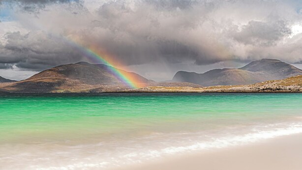 Una spiaggia con acque cristalline turchesi, un arcobaleno e montagne sullo sfondo.