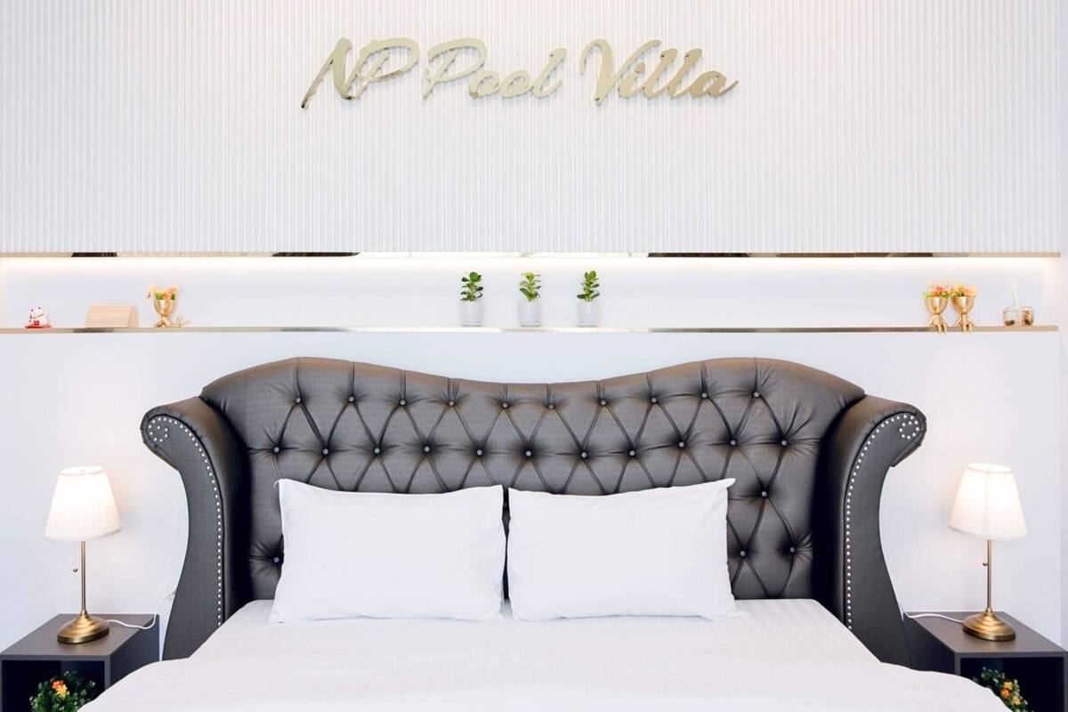 A hotel room with a tufted headboard, white bedding, and a sign reading "NP Pool Villa" on the wall.