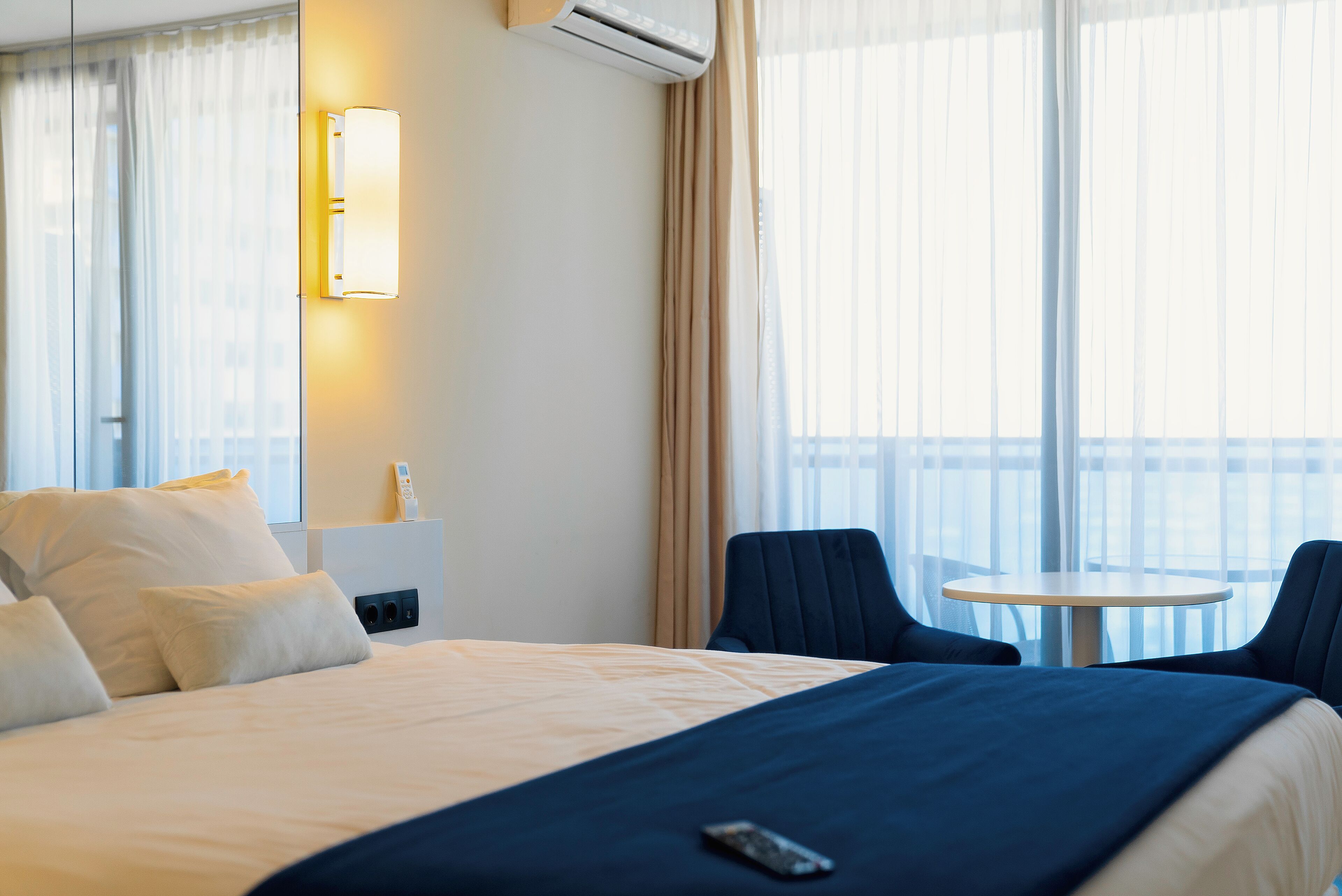 Habitación de hotel con cama, mesita de noche con lámpara, una silla y una pequeña mesa redonda.