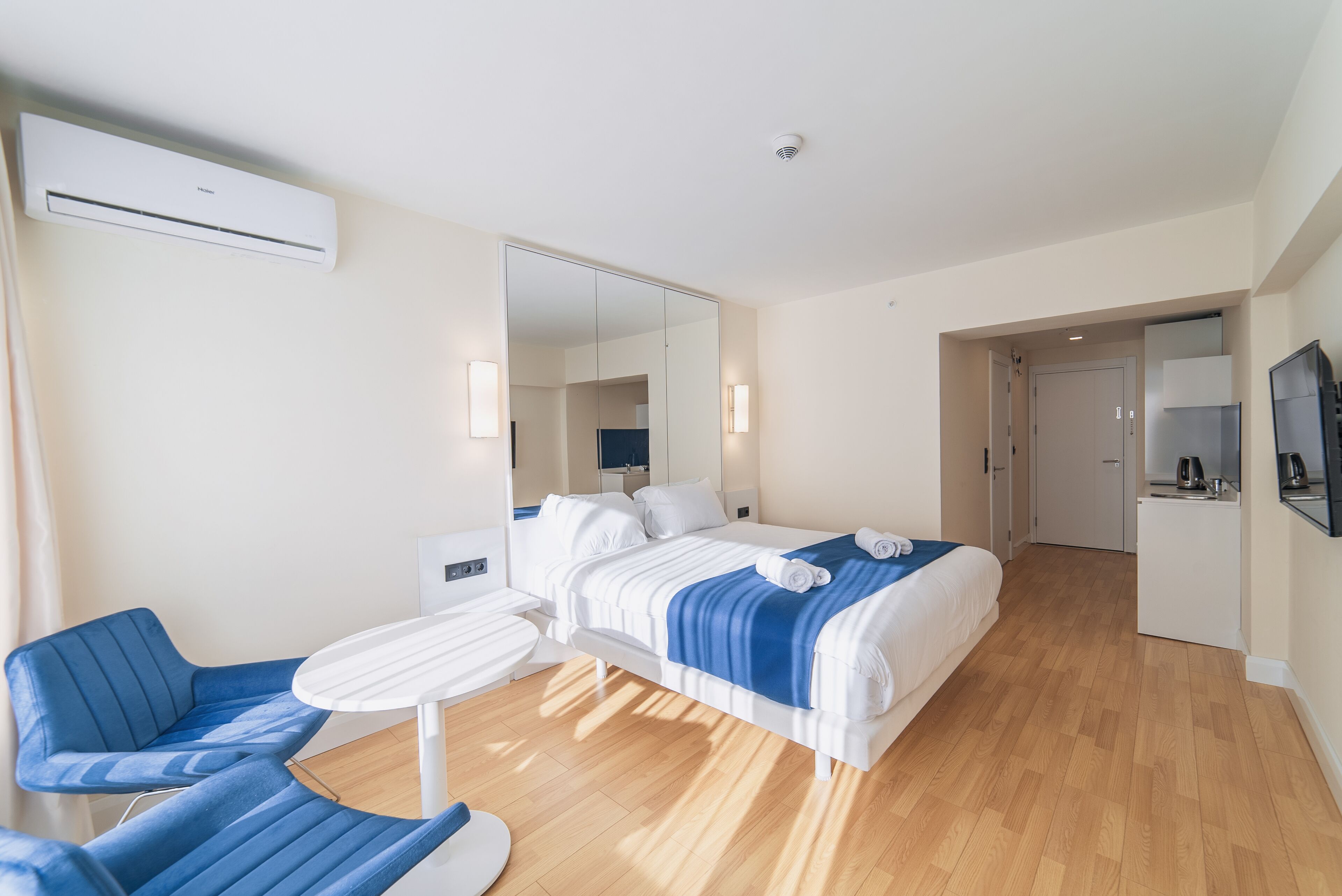 A hotel room with a bed, two blue chairs, a small table, a mirror, and a kitchenette.