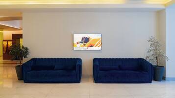 A modern lobby with two blue sofas, a large potted plant, and a framed advertisement on the wall.