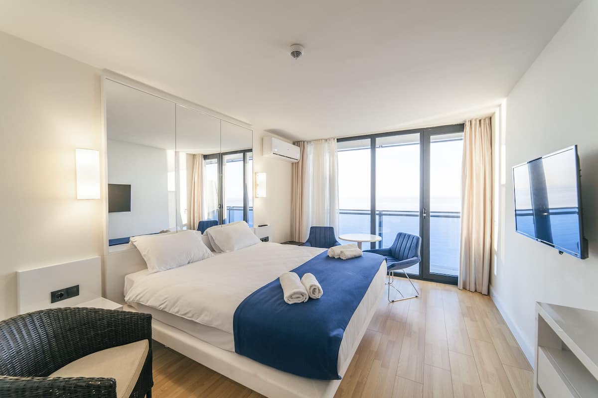 A modern hotel room with a large bed, a flat-screen TV, a balcony with a view, and a dining area.