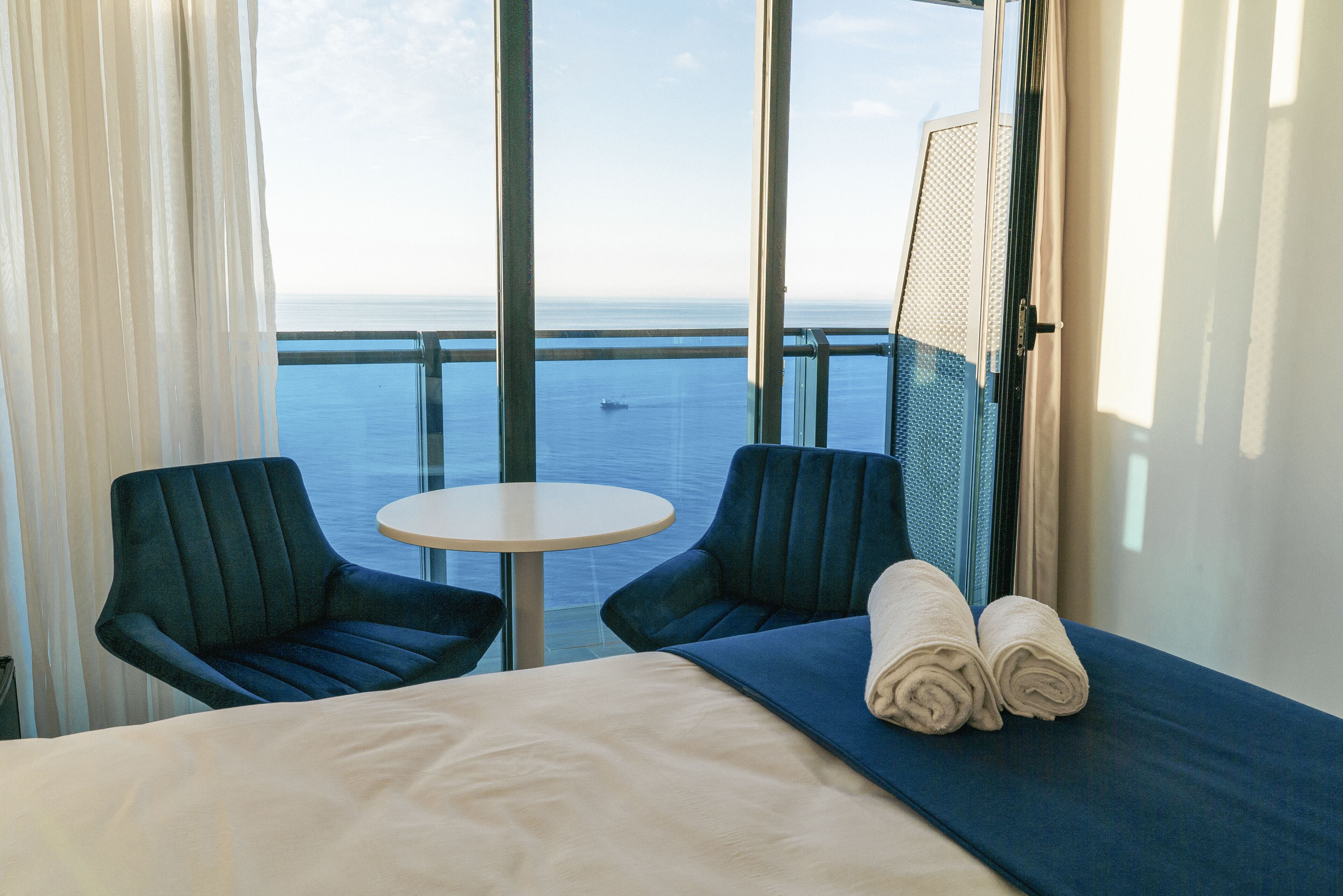 Une chambre d’hôtel avec un lit, deux chaises, une petite table ronde et une vue sur la mer grâce à de grandes fenêtres.
