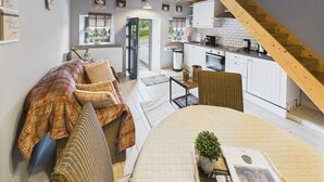 Una sala de estar acogedora con un sofá, una mesa de comedor con una planta y una cocina con armarios blancos y una escalera de madera.