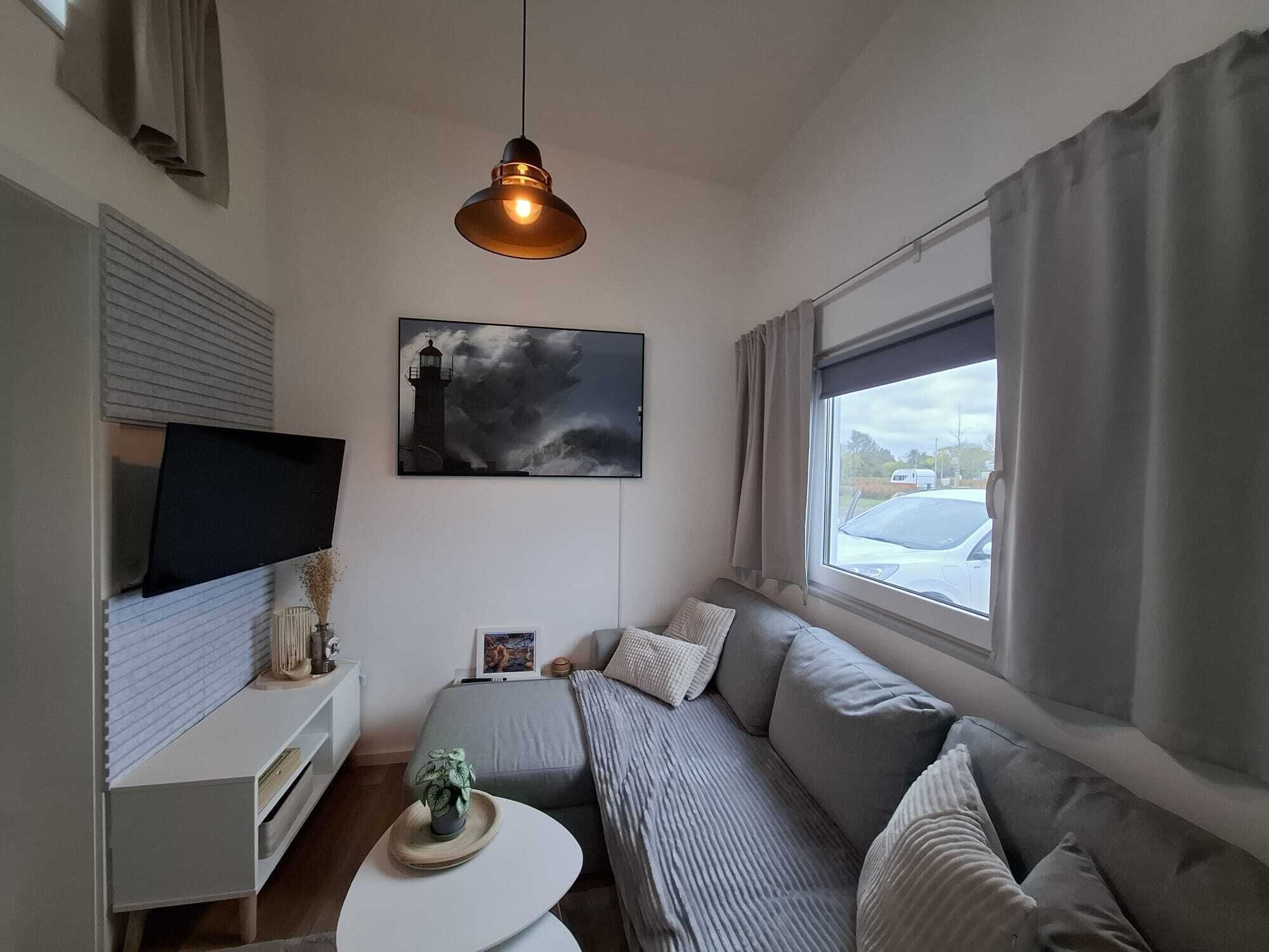 Un salon moderne avec un canapé gris, une petite table blanche, un téléviseur posé sur un meuble blanc, un tableau représentant un phare et une fenêtre avec des rideaux.