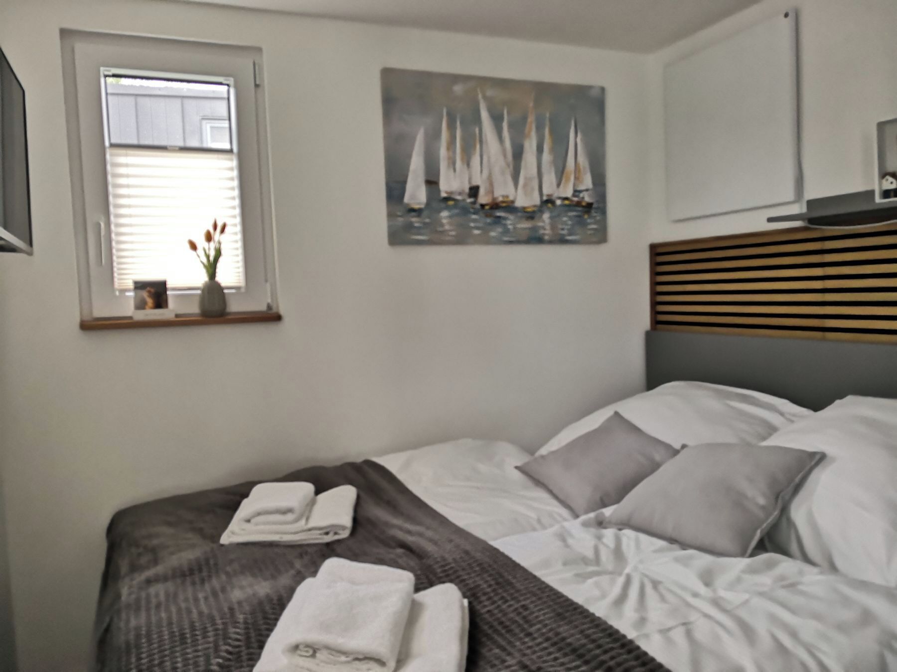 A neatly made bed with pillows and a folded towel, a window with a view of the outside, and a painting of sailboats on the wall.