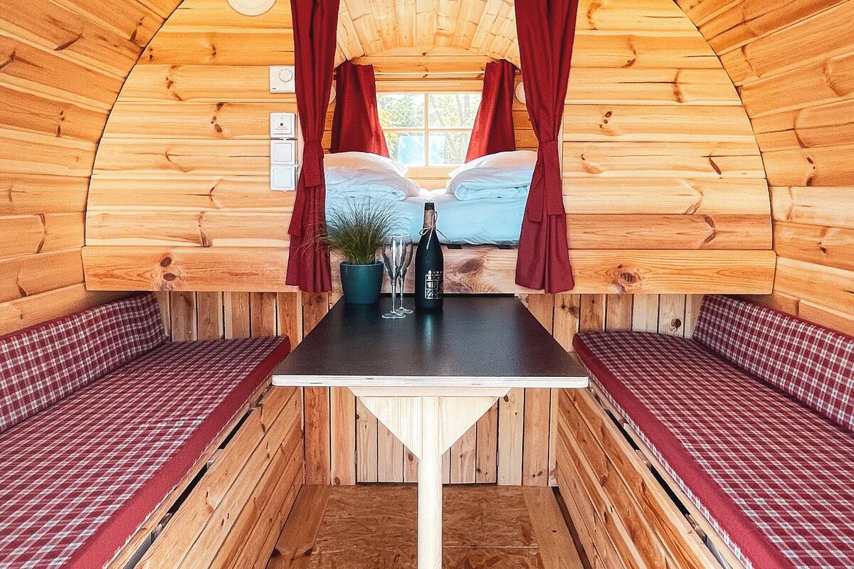 A cozy cabin interior with a wooden interior, a bed with red curtains, a table with a bottle and glasses, and checkered benches.