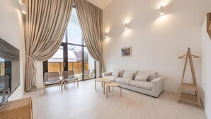 A modern living room with a white sofa, two armchairs, a small round table, and a wooden ladder. Large windows with beige curtains allow natural light.