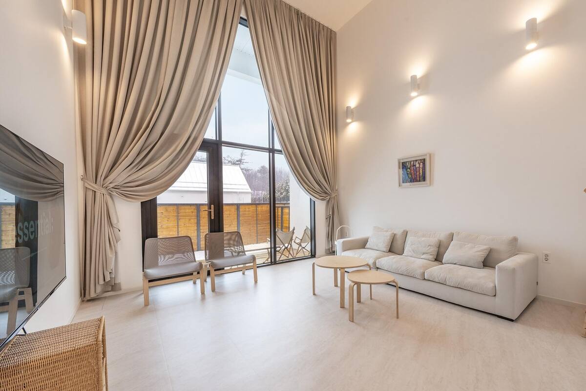 A modern living room with a white sofa, two armchairs, a small round table, and a wooden ladder. Large windows with beige curtains allow natural light.