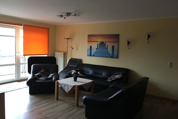 Un salon avec un canapé en cuir noir, une petite table sur laquelle est posé un vase, un tableau représentant une jetée et un ventilateur de plafond.