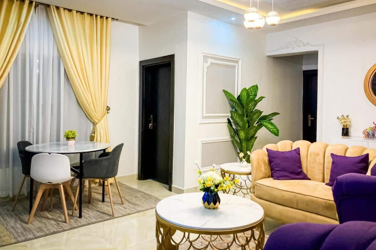 Una sala de estar moderna con una mesa redonda, un sofá color beige con cojines morados, una mesa de comedor con sillas y una planta grande de color verde.