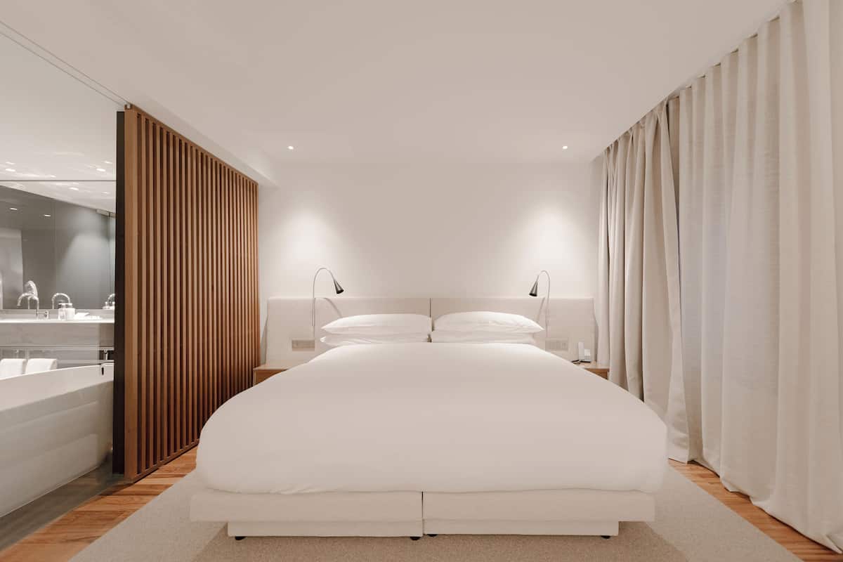A modern bedroom with a large bed, bedside lamps, a wooden panel wall, and a bathroom visible to the left.
