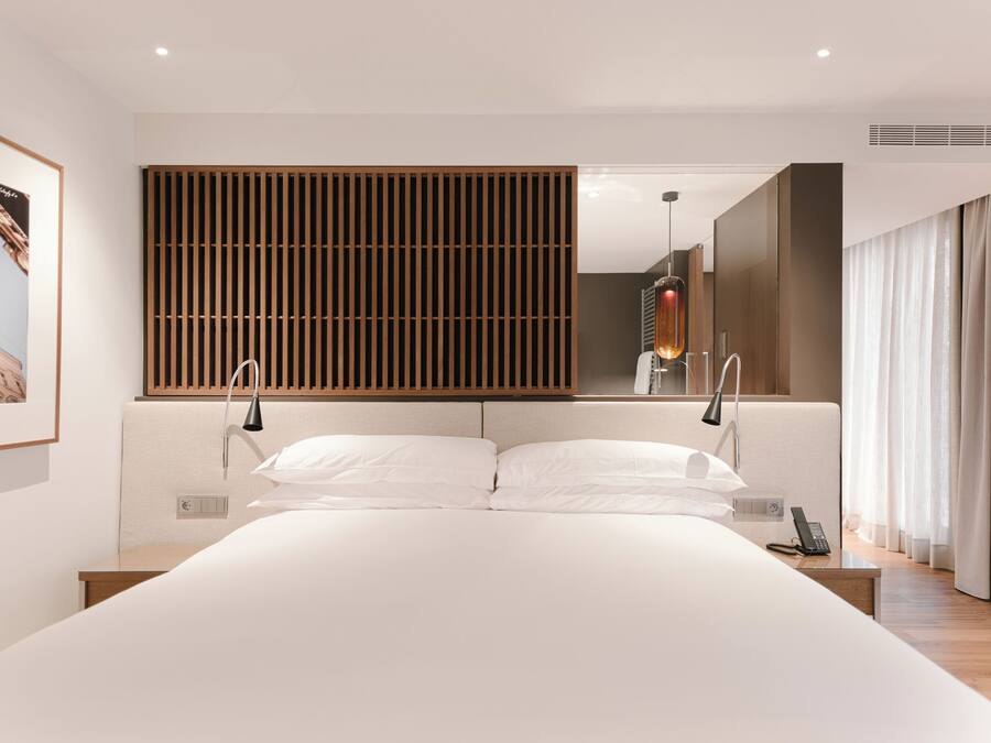 A modern hotel room with a large bed, a wooden slat headboard, a bedside table with a lamp, and a large mirror.