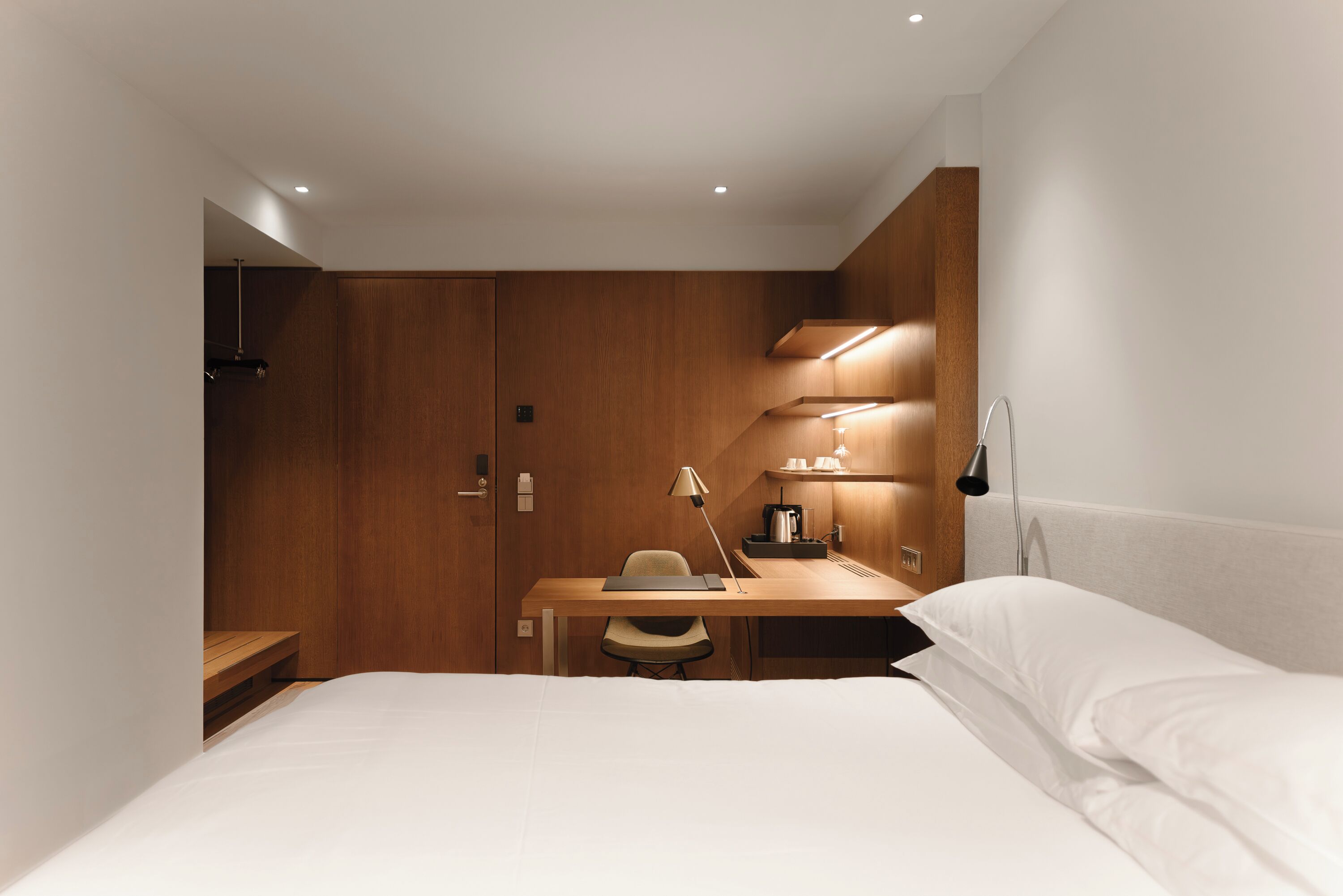 Quarto de hotel compacto com cama, escrivaninha, cadeira, luminária e pequena área de cozinha.