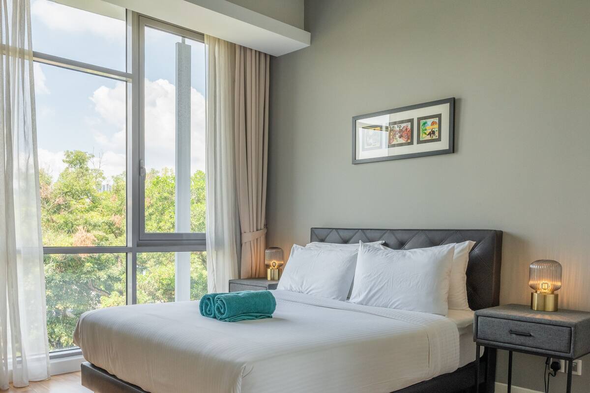 Une chambre d’hôtel avec une grande fenêtre, un lit avec du linge de maison blanc, une table de chevet avec une lampe et un tableau encadré au mur.