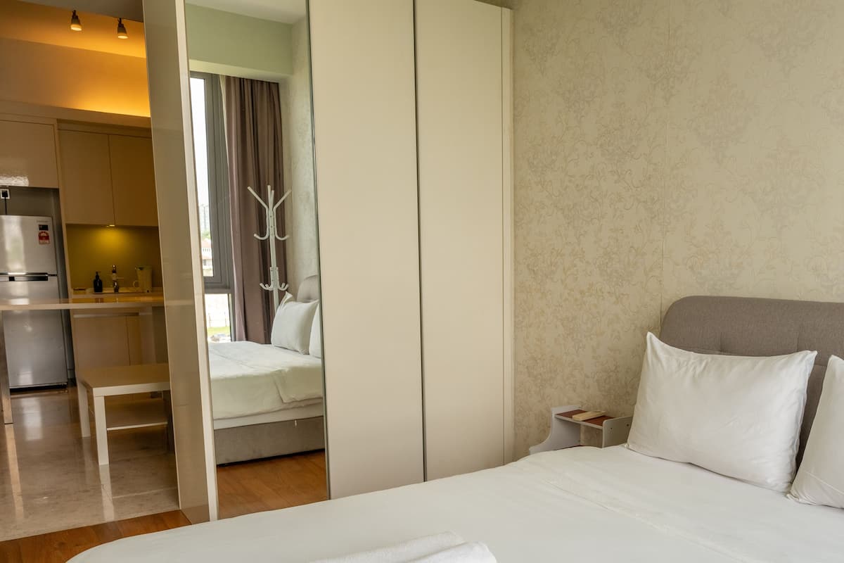 Une chambre d’hôtel avec un lit, une table de chevet, un miroir, une armoire et, au fond, une petite cuisine.