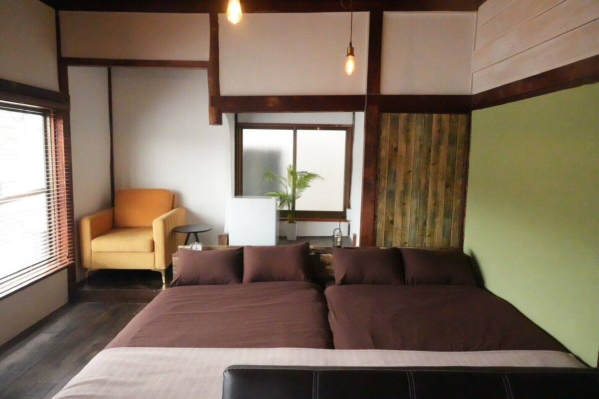 A room with a bed, a chair, a small table, and a window with blinds.