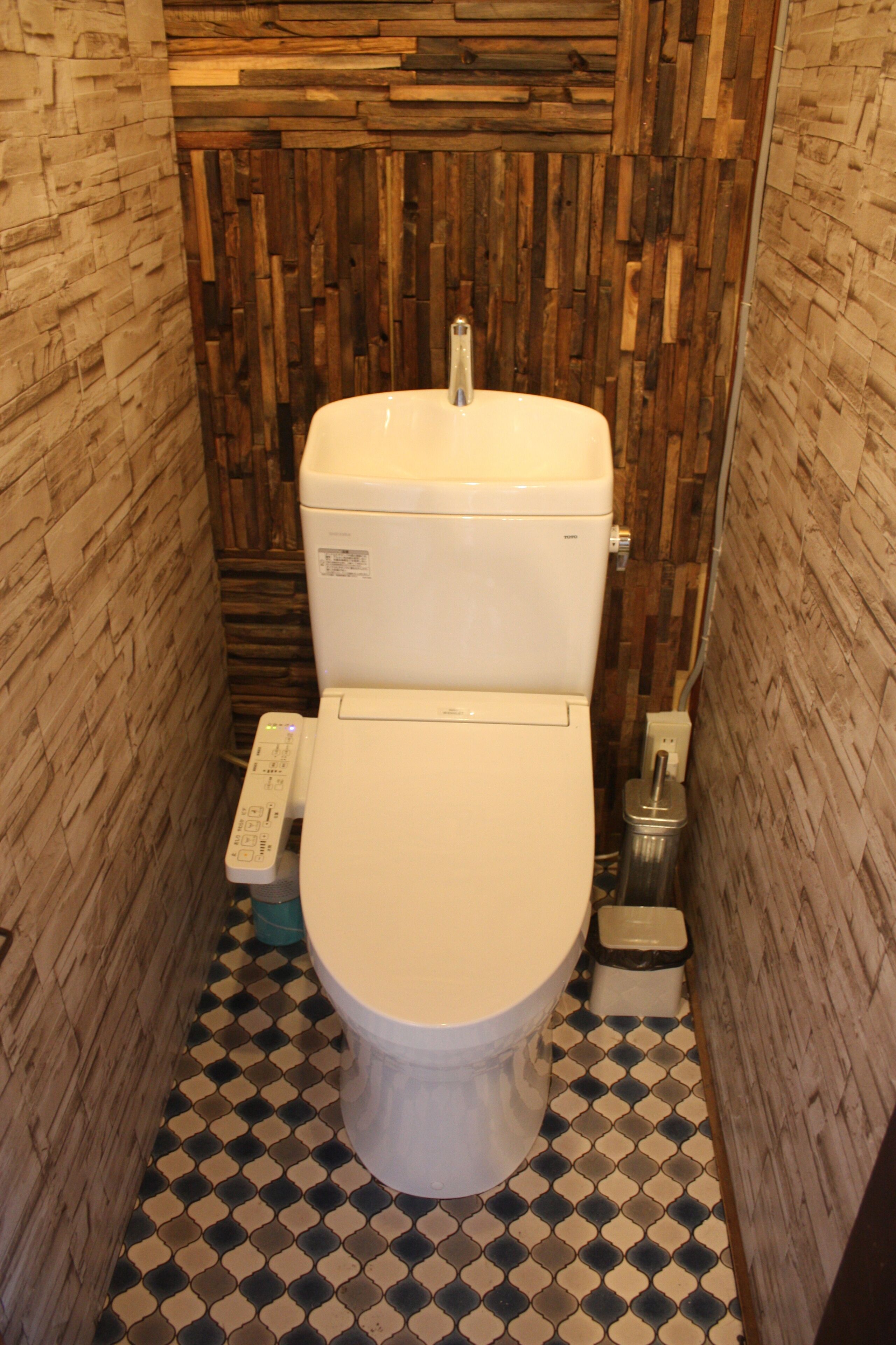 A compact bathroom with a toilet, a bidet, and a hand dryer. The walls are covered with wooden panels, and the floor has a checkered tile pattern.