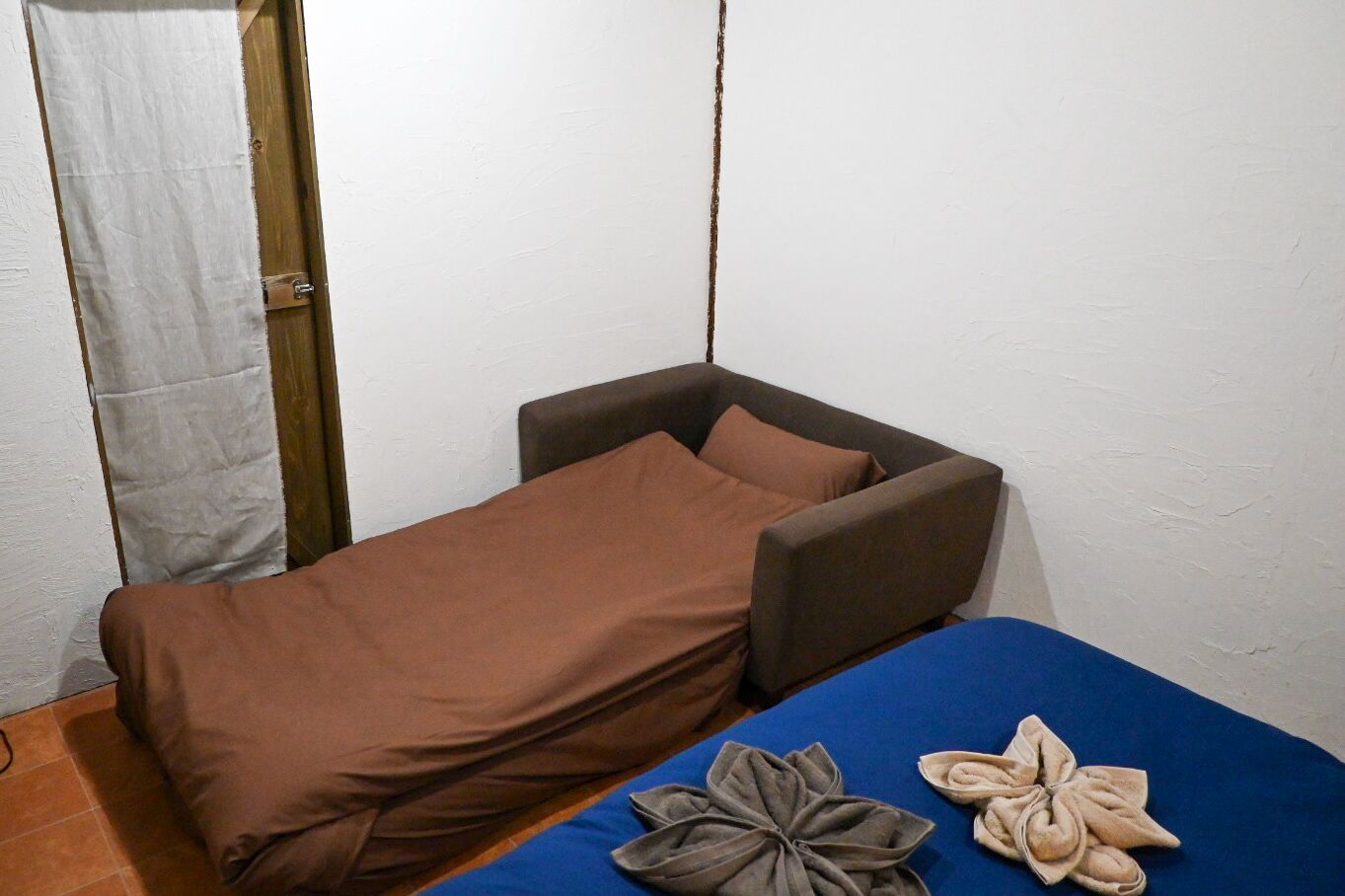 A small, single-bed room with a brown bedspread and pillows, a blue blanket, and a decorative towel on the floor.