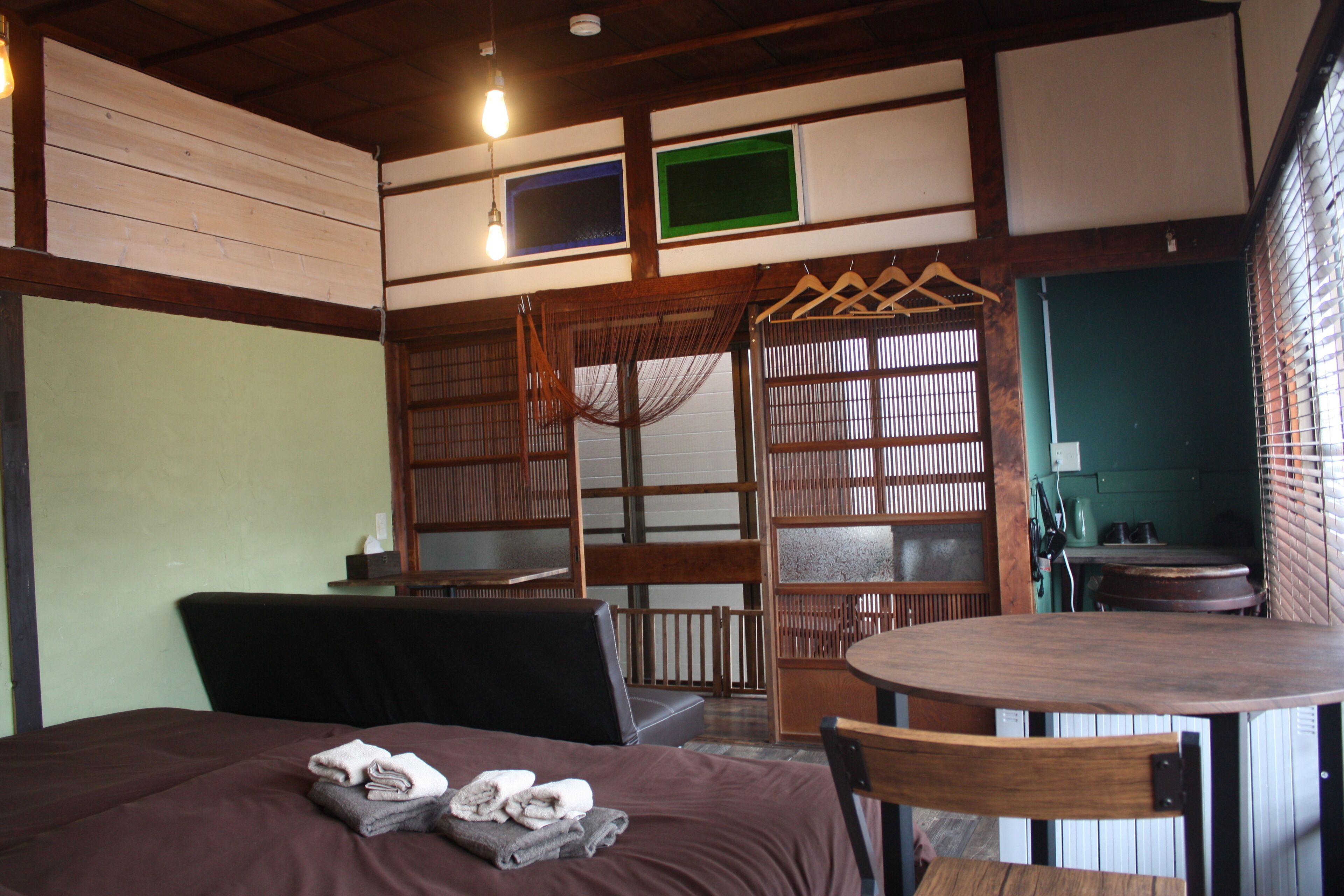 A room with a bed, a table, and a chair. There is a hanging rack with clothes and a small table with a kettle.