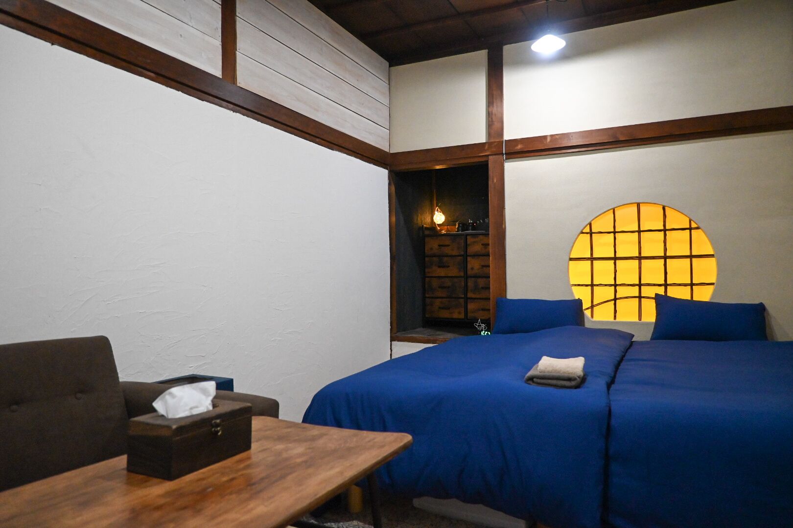 Une pièce comprenant un lit, une table en bois avec une boîte de mouchoirs, un canapé et un mur percé d’une fenêtre ronde.