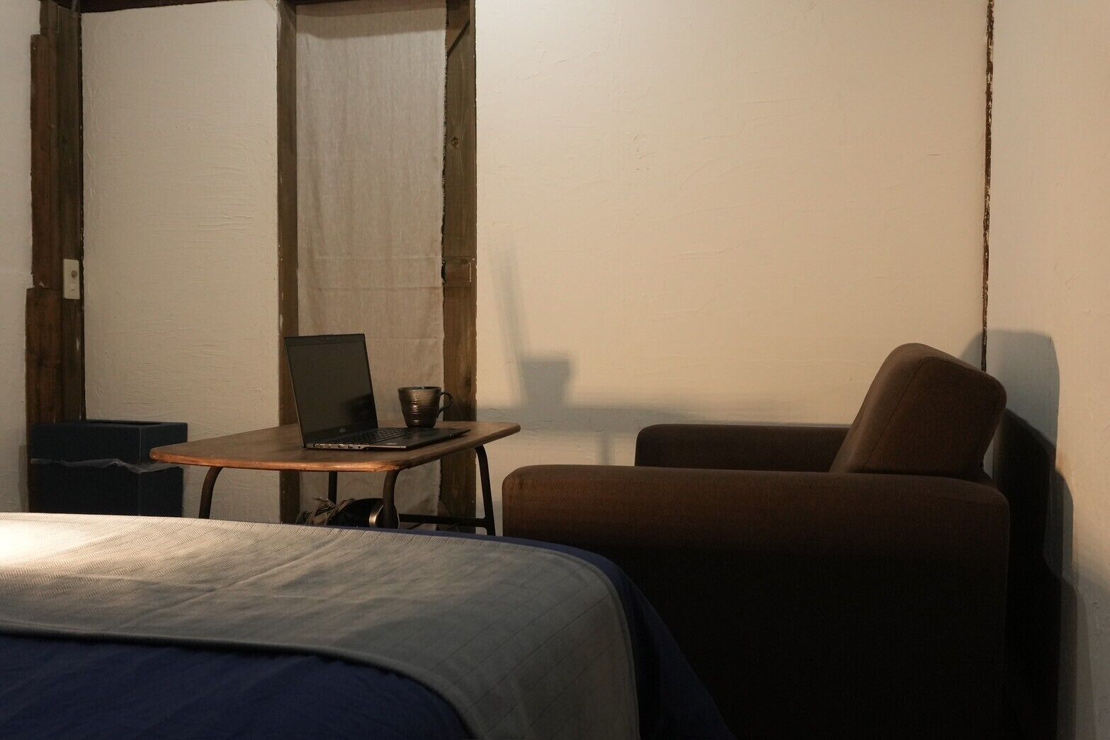 A small, dimly lit room with a single bed, a chair, and a table with a laptop and a cup.