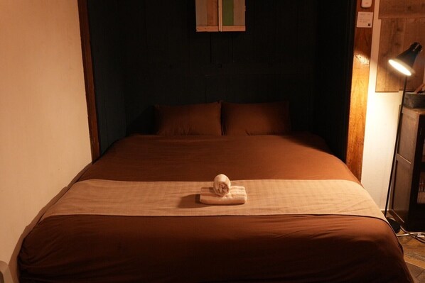 A neatly made bed with a rolled towel in the center, a wooden headboard, and a small table lamp to the right.