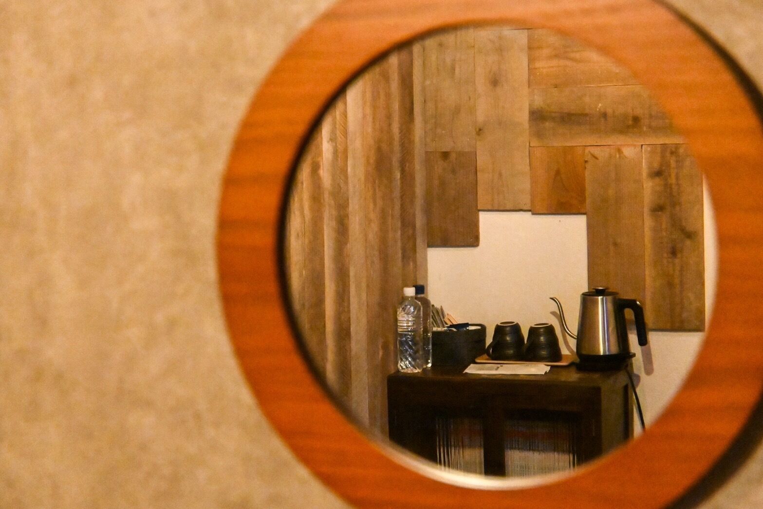 A circular mirror reflecting a wooden wall, a table with a kettle, cups, and a bottle.