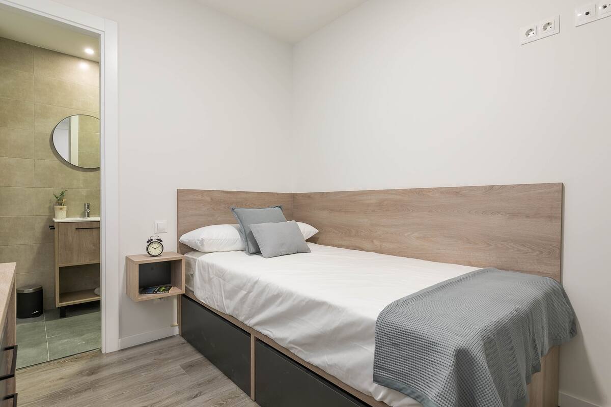 A modern bedroom with a wooden headboard, a single bed with a grey blanket, a bedside table with a clock, and a small shelf.