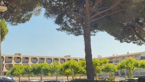 A parking area with a tree, a building in the background, and a blue sign on the dashboard. - Studio Proche Centre ET Plage Avec Clim ET Parking (La Grande-Motte)