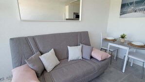 A grey sofa with multiple cushions, a small dining table with chairs, and a wall-mounted mirror. - Studio Proche Centre ET Plage Avec Clim ET Parking (La Grande-Motte)