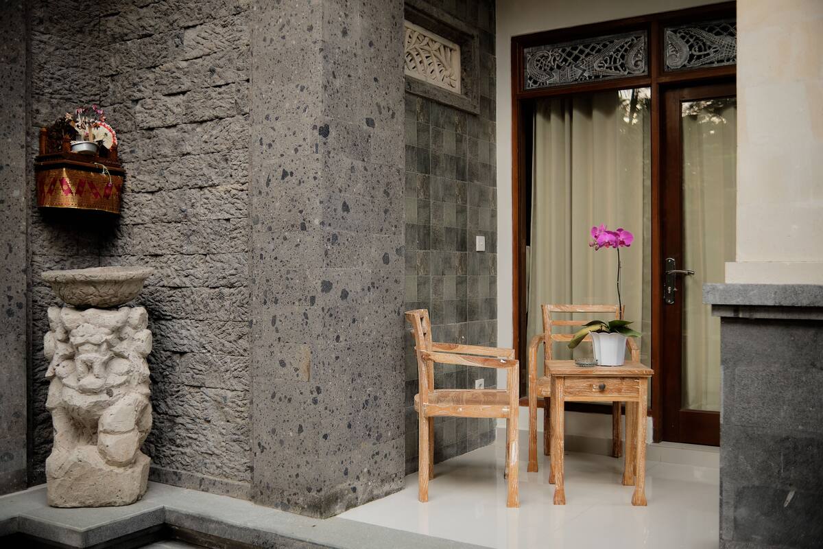 A stone pillar, two wooden chairs, and a potted plant near a door with a decorative window.