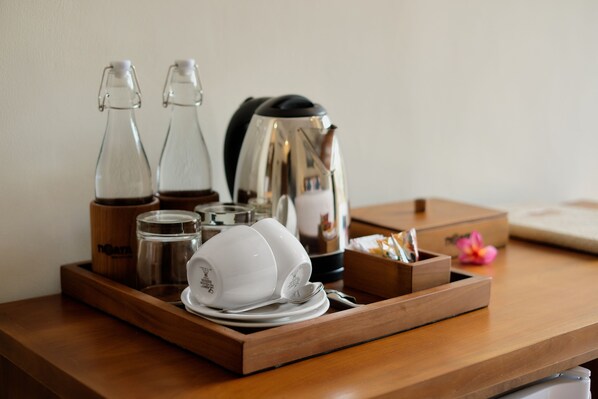 A wooden tray with a coffee maker, two glass bottles, a white teapot, and a small flower on a table.