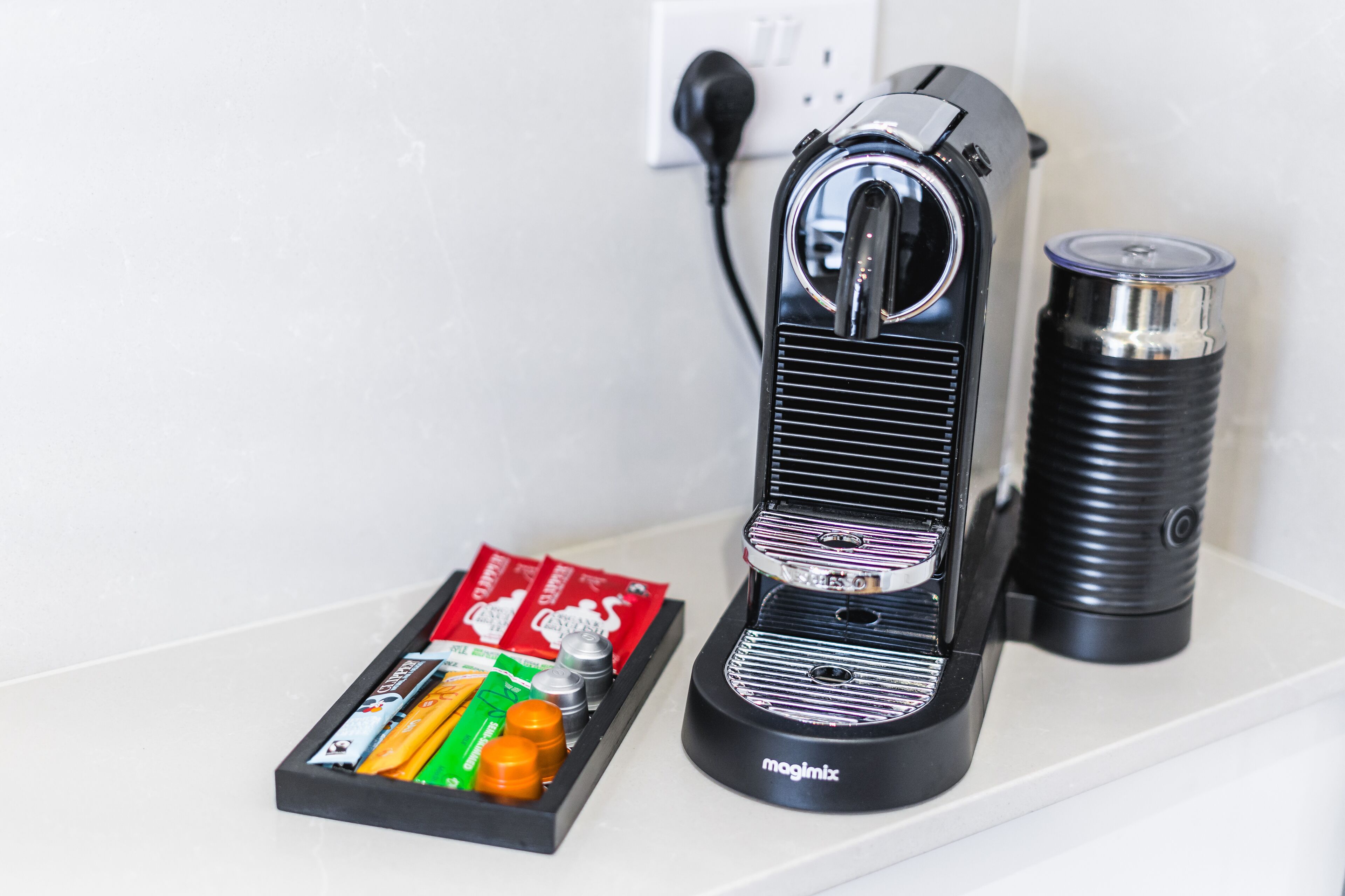 A Magimix coffee machine with a black base and a transparent water tank, placed on a white surface next to a power outlet.