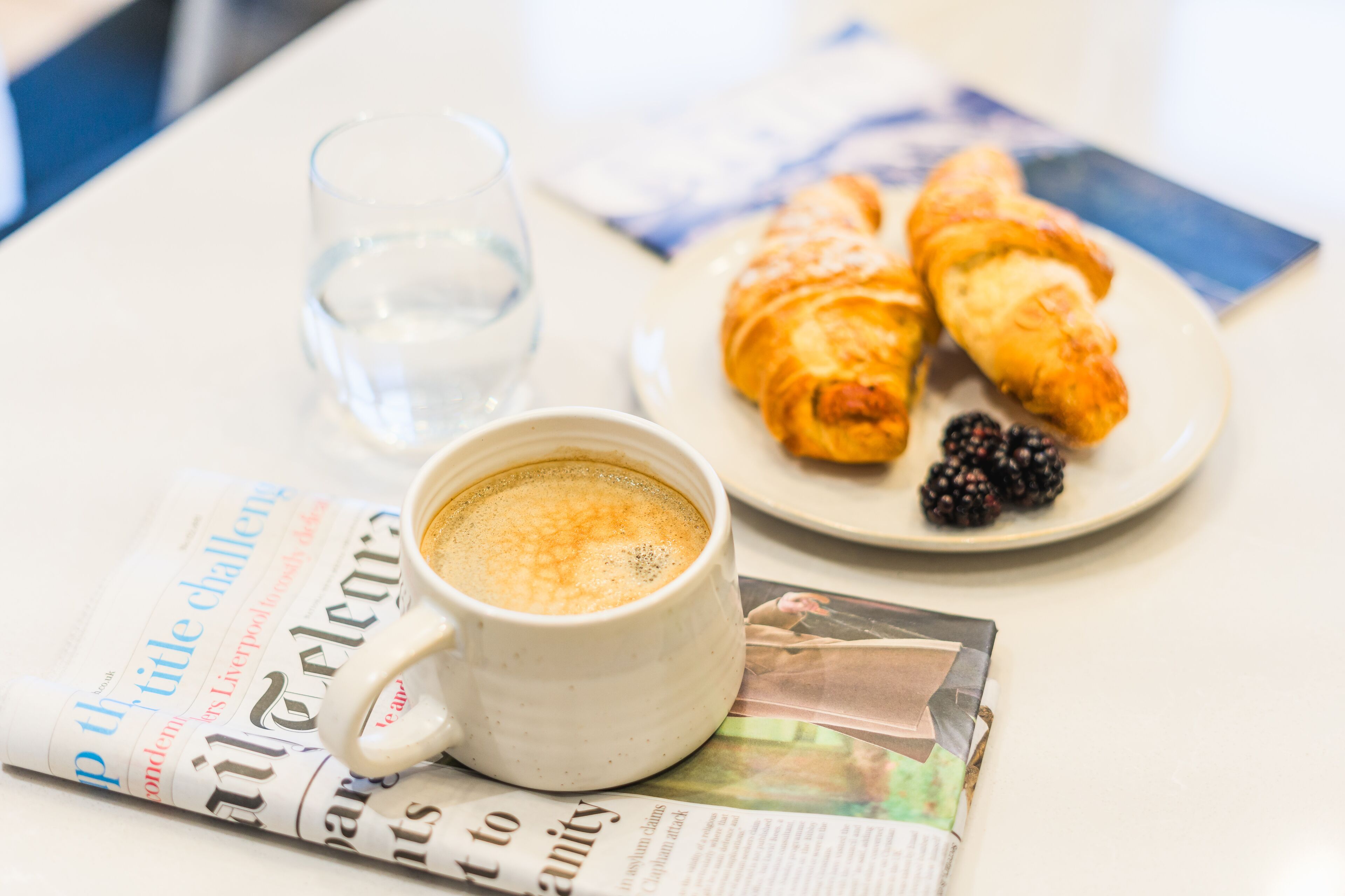 A cup of coffee, a glass of water, and a plate with croissants and blackberries on a table with a newspaper.
