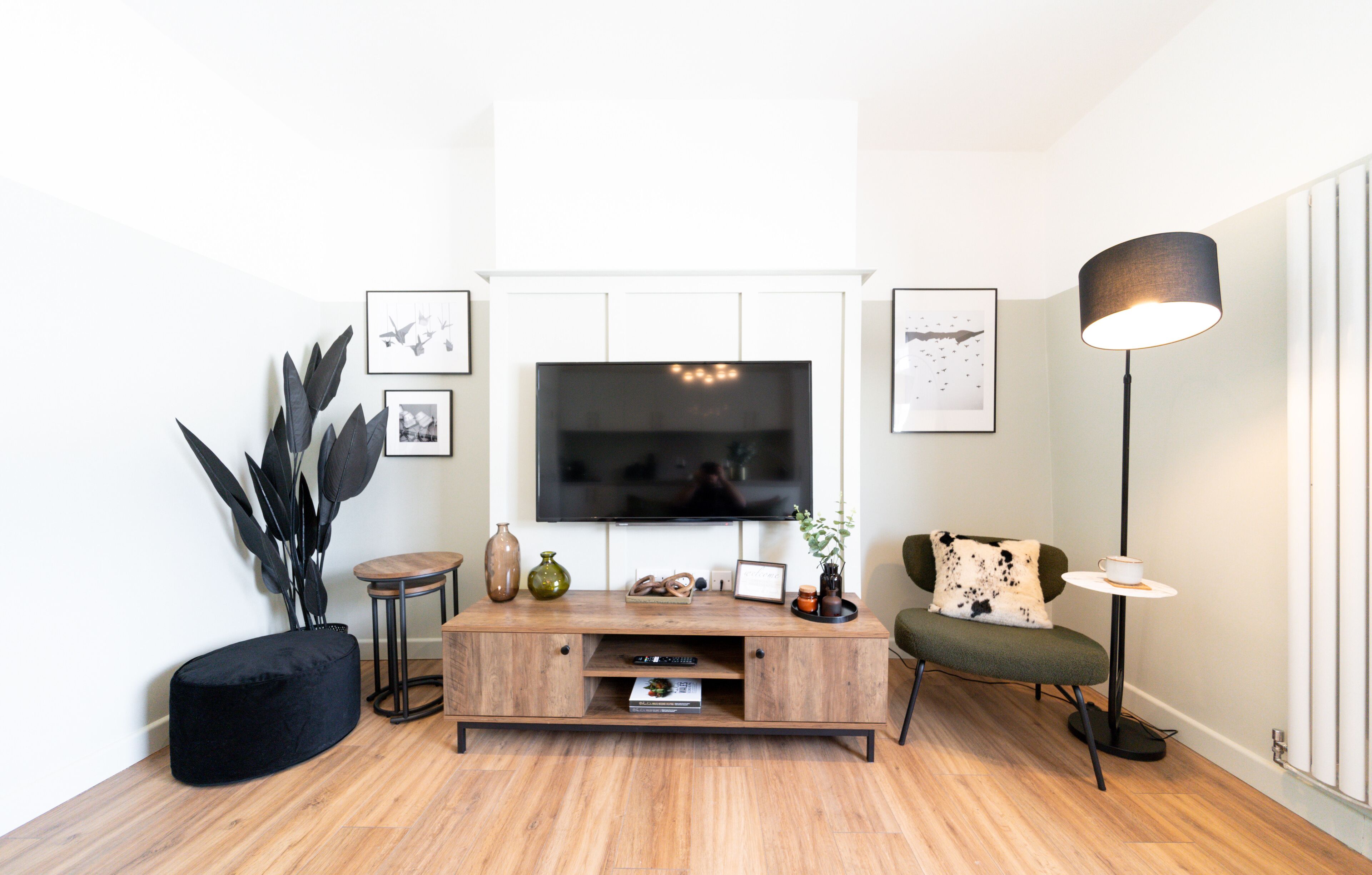 Un salon moderne doté d’un téléviseur à écran plat, d’un meuble de rangement en bois, d’un pouf noir, d’un fauteuil vert, d’une lampe sur pied et d’œuvres d’art murales.