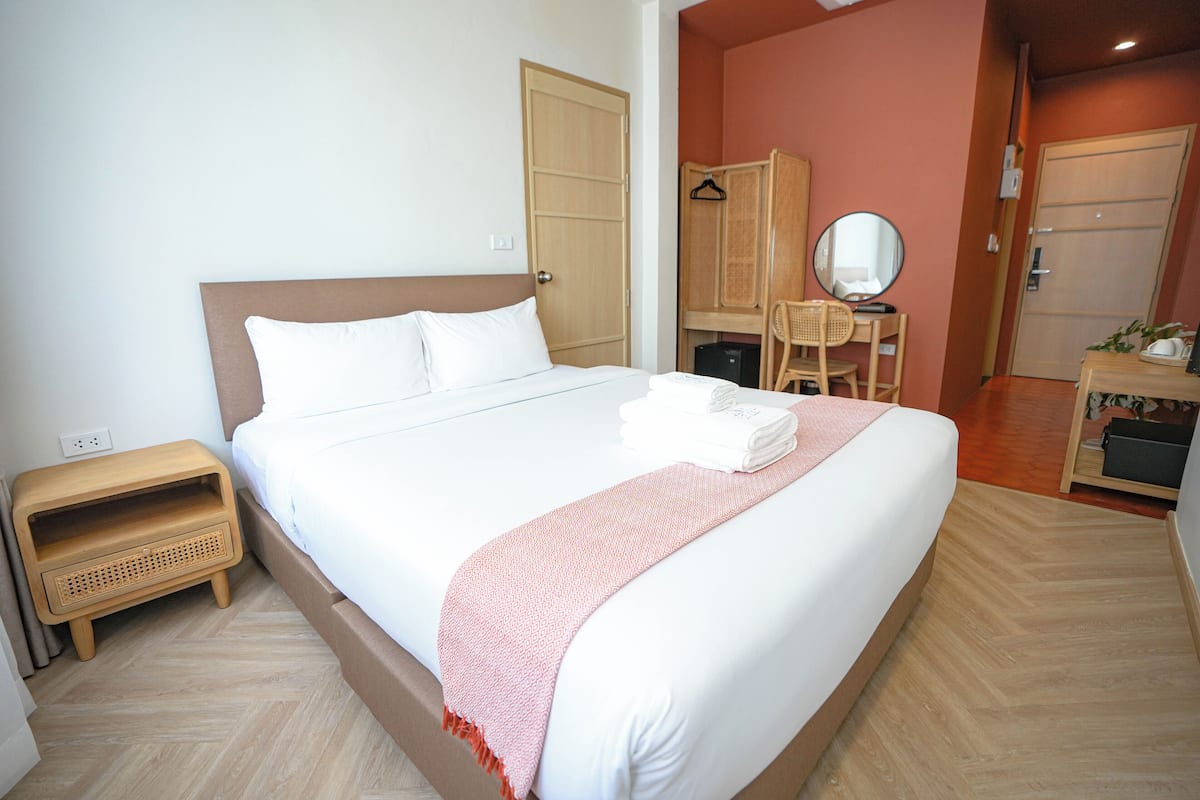 Un lit bien fait, avec une couette blanche et un plaid rose, une table de chevet en bois surmontée d’un panier en osier, une armoire en bois et un miroir accroché au mur.